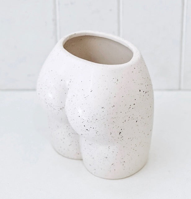 A beige ceramic vase with a playful, body-shaped design, resembling a bum, placed against a white tiled background.